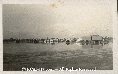 An unlabeled photograph of a flooded settlement, low buildings and palm trees reflected in standing water