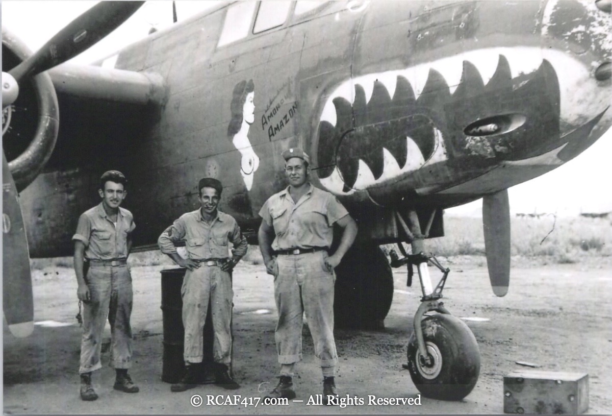 Nose art reading Amoroso Amazon, pin-up figure beside a large shark-mouth nose, with three crew members
